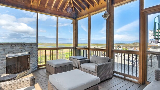 A covered outdoor deck with wicker furniture, a stone fireplace, and a view of the landscape. - Summer Salt: Amazing Ocean, Marsh and Lighthouse Views (Caswell Beach)