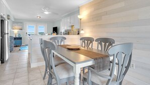 A modern kitchen with a dining area, featuring a long table with chairs, a wooden backsplash, and a view of the outdoors. - Summer Salt: Amazing Ocean, Marsh and Lighthouse Views (Caswell Beach)