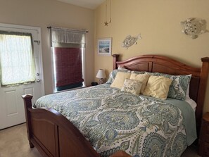 A bedroom with a wooden bed, patterned bedding, a window with curtains, a bedside lamp, and wall decorations. - The Marlin at Gulf Stream invites you to relax with an Ocean Breeze. (Myrtle Beach)
