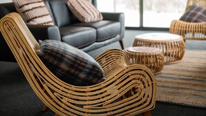 A wicker chair with a plaid cushion, a round ottoman, and a dark leather sofa in a living room with a large window.