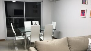 Una sala de estar moderna con un sofá color crema, una mesa de centro de vidrio y sillas blancas. Hay un ventanal con persianas, un equipo de aire acondicionado en la pared y un televisor de pantalla plana montado en la pared.