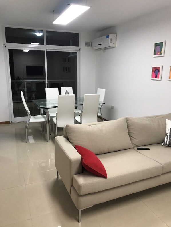 A modern living room with a beige sofa, a glass coffee table, and white chairs. There is a large window with blinds, an air conditioning unit on the wall, and a flat-screen TV mounted above the TV.