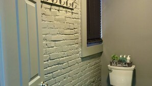 Un baño con pared de ladrillo, un inodoro, un cesto de basura y una ventana pequeña.