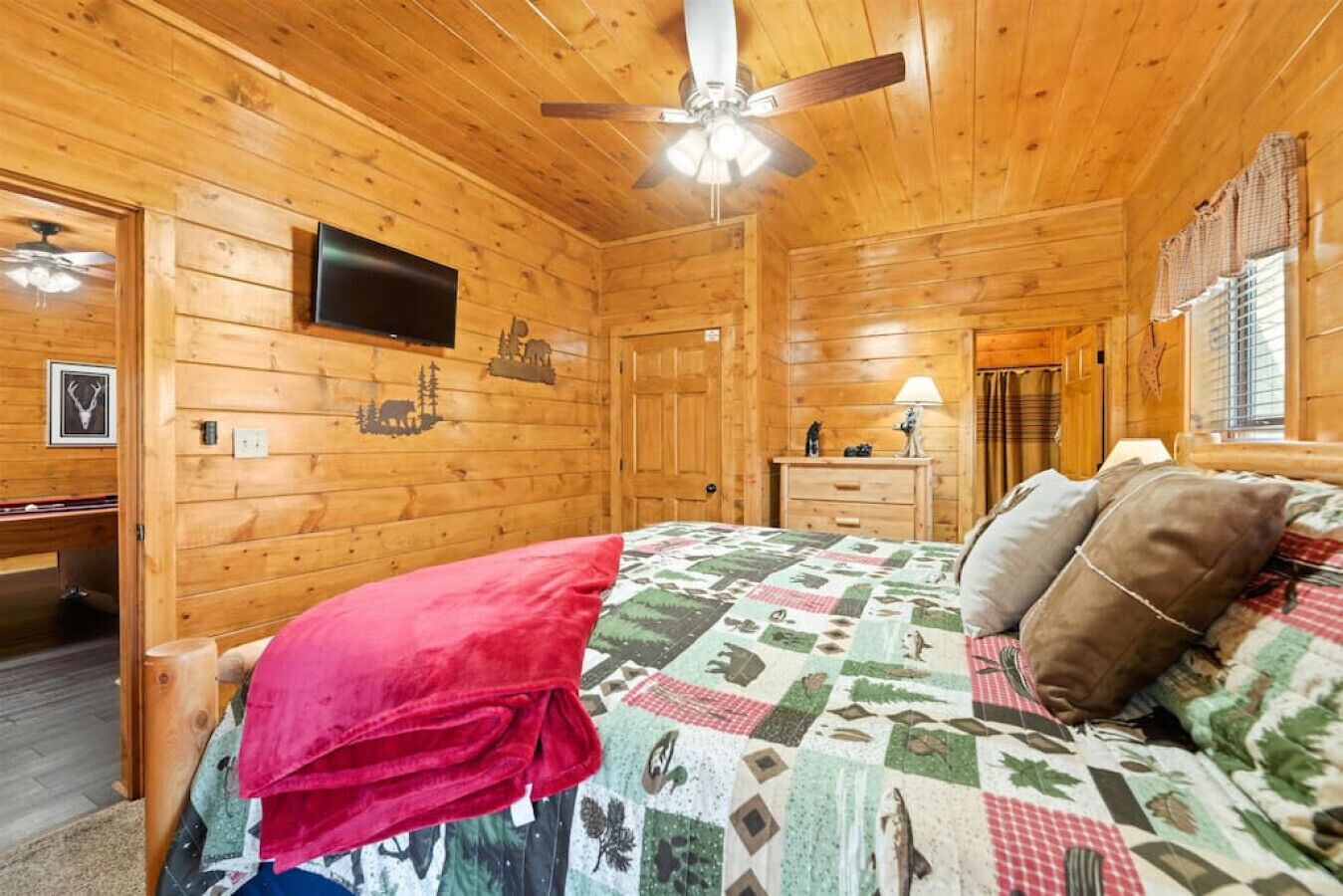 A wooden cabin bedroom with a bed, a dresser, a television, and a ceiling fan.