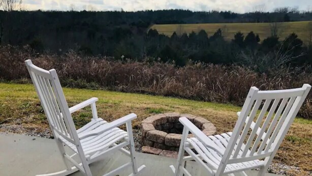 Two white rocking chairs facing a fire pit on a concrete patio with a view of a field and forest in the background. - Bourbon Country Cottage <br>Newly renovated  in the country with amazing views (Mount Eden)