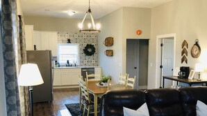 A living room with a black leather sofa, a dining table with chairs, a kitchen in the background, and a chandelier. - Bourbon Country Cottage <br>Newly renovated  in the country with amazing views (Mount Eden)