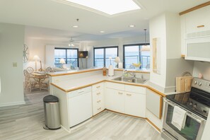 A modern kitchen with white cabinetry, a dishwasher, and an oven. There is a dining area with a table and chairs, and a view of the ocean through large windows.