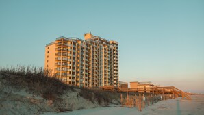 Un hotel de gran altura con varios balcones, situado cerca de una playa con un paseo de madera que lleva al agua.