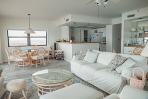 A modern living room with a white sofa, round glass coffee table, wicker chairs, and a dining area with a glass table.