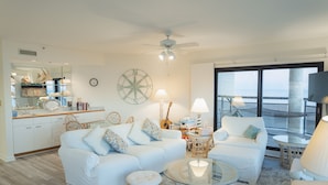 Amplia sala de estar con sofá blanco, mesa de centro de vidrio y silla de mimbre. Se observa también un ventilador de techo y un amplio ventanal con vistas al exterior.