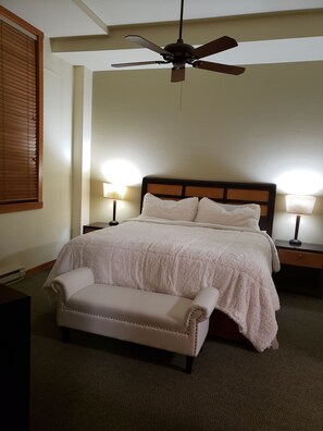 A bedroom with a bed, two bedside lamps, a ceiling fan, a bench, and a window with blinds.