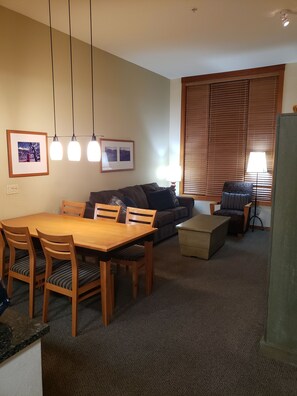 A living room with a wooden dining table, chairs, a sofa, and a window with blinds.