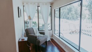 Ein Zimmer mit großem Fenster, einem Sessel, einem kleinen Tisch mit Lampe und Blick auf einen Zaun und Bäume.