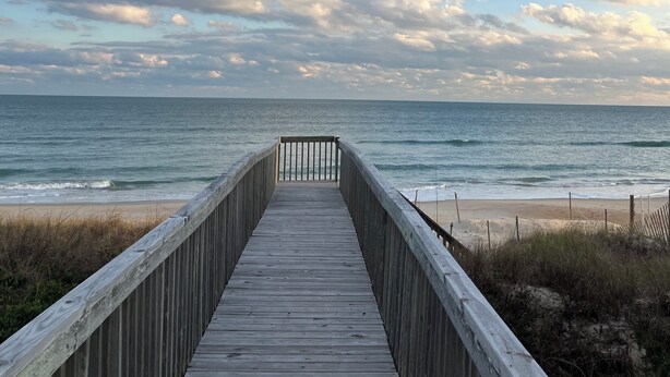 A wooden boardwalk leading to a beach with a clear view of the ocean and a partly cloudy sky.