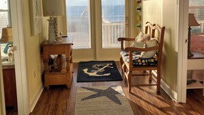 A cozy room with wooden flooring, a rocking chair, a small table with a lamp, a rug with an anchor design, and a view through a door.