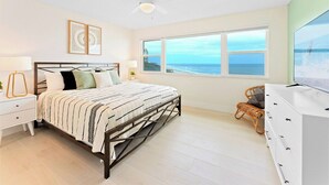 A bedroom with a bed, bedside tables, a dresser, a ceiling fan, and a large window with a view of the ocean. - Gorgeous Oceanside Oasis w/ Heated Pool, Oceanfront Patio & Steps to the Beach 🏖️☀️ (Satellite Beach)