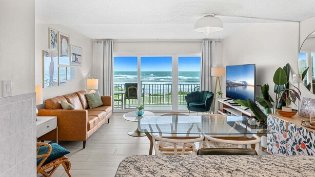 A modern living room with a large window overlooking the ocean, a glass dining table, and comfortable seating. - Gorgeous Oceanside Oasis w/ Heated Pool, Oceanfront Patio & Steps to the Beach 🏖️☀️ (Satellite Beach)