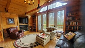 Interior acollidor d'una cabana amb un sofà de cuir, una llar de foc, una biblioteca i una finestra gran.