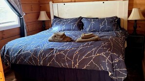 Un dormitori de cabana de fusta amb un llit, dues làmpades de nit, un ventilador de sostre i una finestra amb cortines.