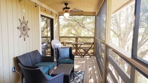 A covered porch with wicker furniture, a ceiling fan, and a view of trees through large windows.