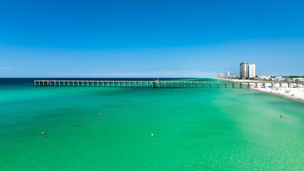 A coastal scene with a pier extending into turquoise waters, a beach with people, and a cityscape in the background.