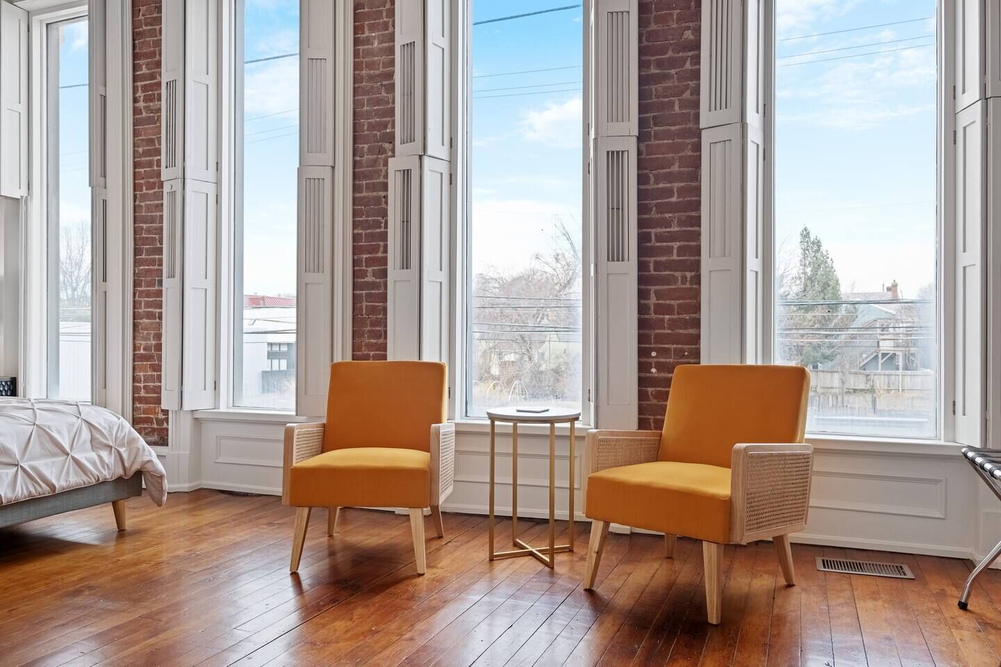 Quarto com cama, duas poltronas, uma mesinha e janelas amplas com vista para edifícios.