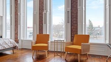 Quarto com cama, duas poltronas, uma mesinha e janelas amplas com vista para edifícios.