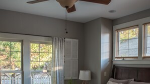 A bedroom with a bed, a ceiling fan, a window with blinds, a nightstand with a lamp, and a view of a fenced outdoor area.