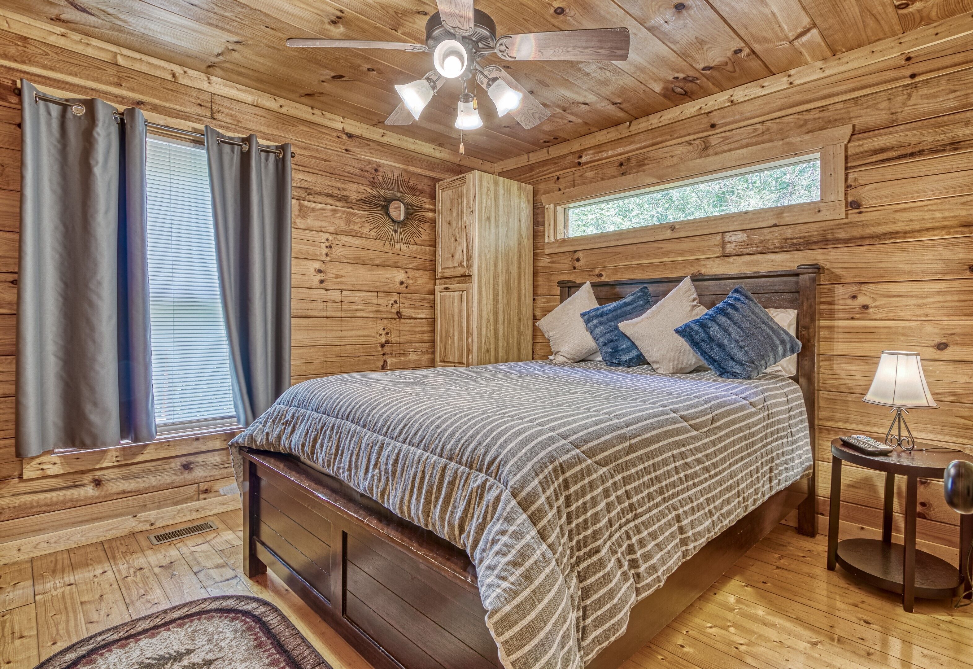 A wooden cabin bedroom with a bed, curtains, a ceiling fan, a window, and a nightstand.