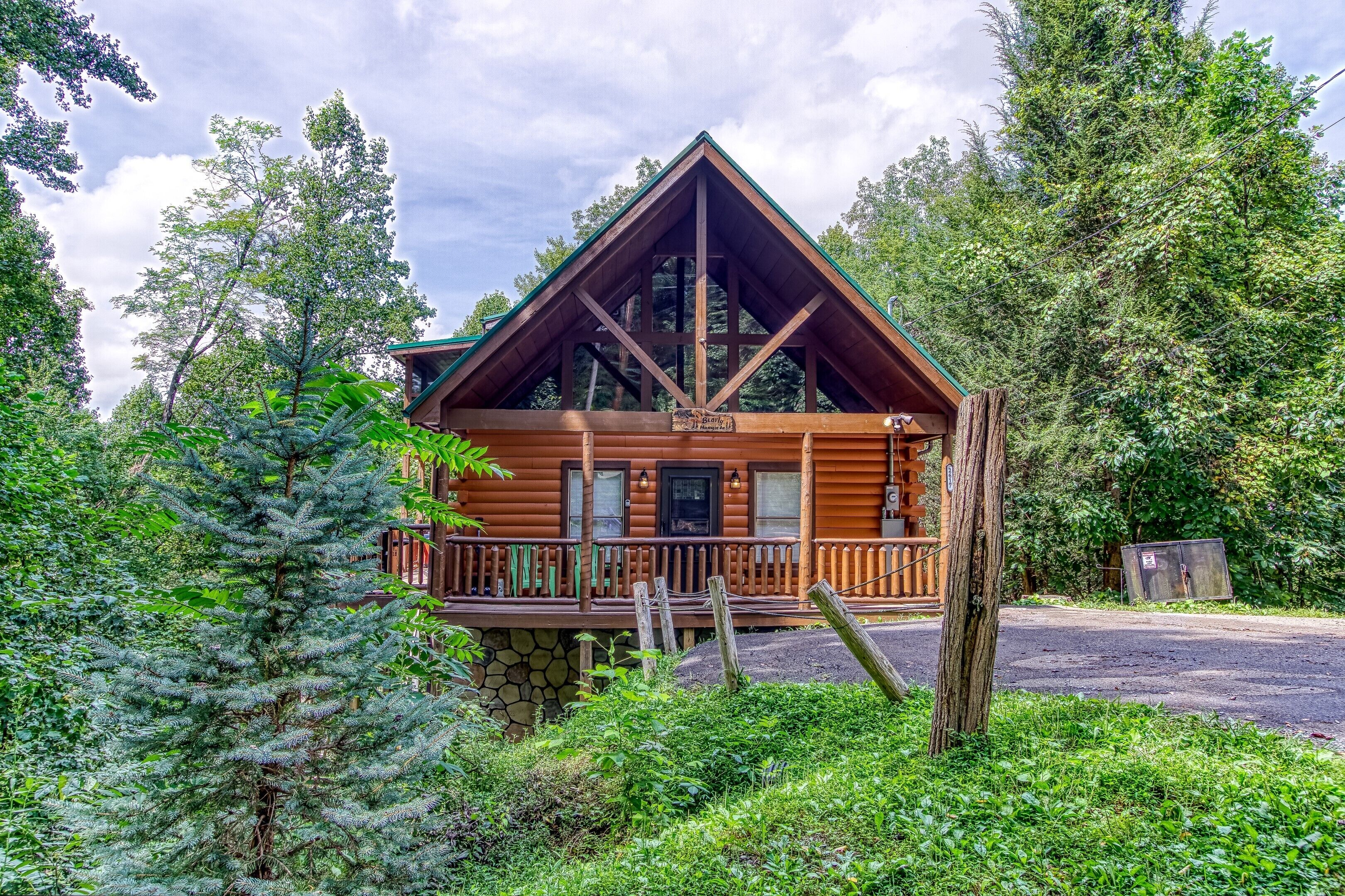 A log cabin with a wooden deck and a stone foundation, surrounded by dense forest.