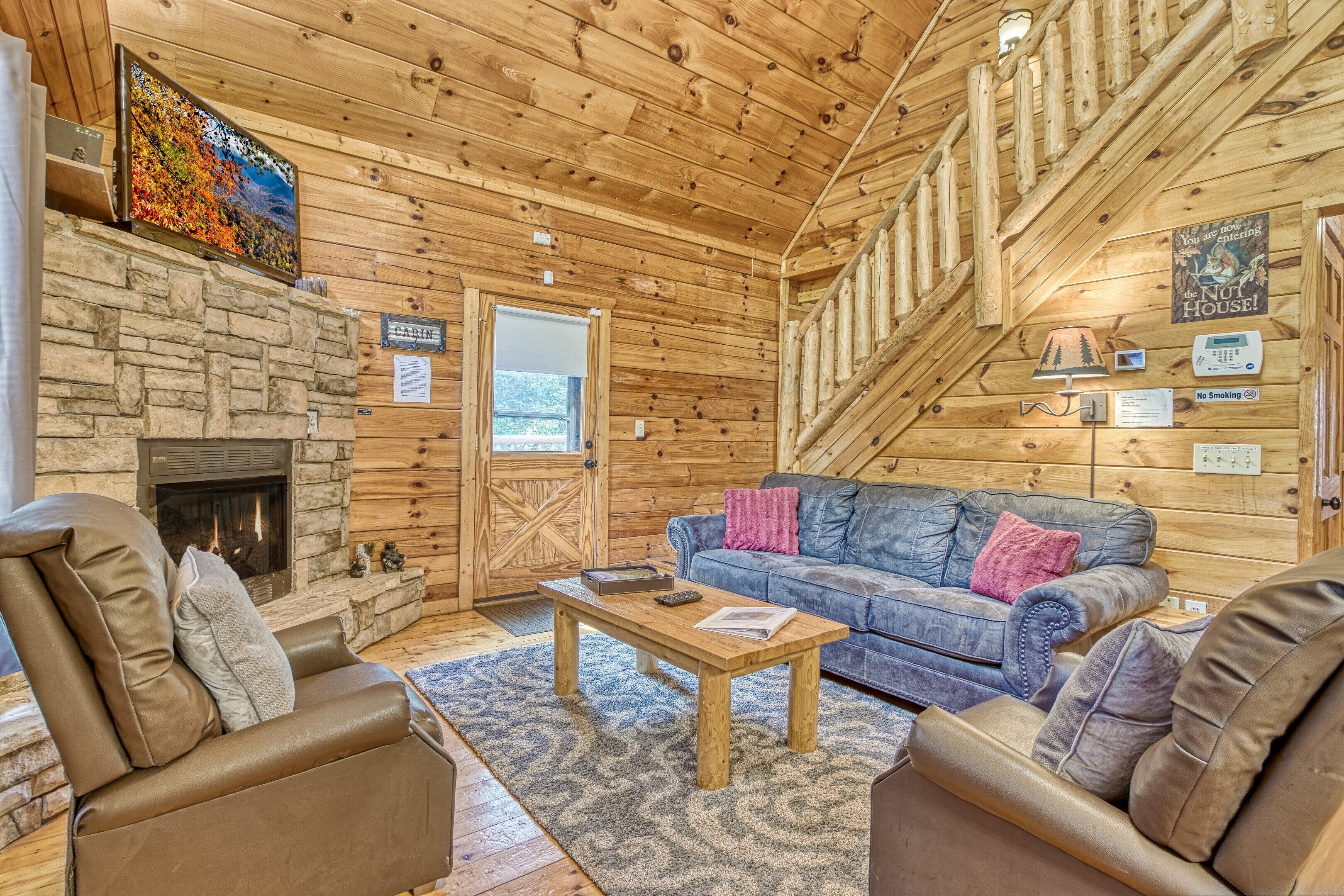 A cabin interior with a stone fireplace, a blue sofa, and a wooden staircase.