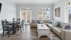 A spacious living area with a dining table, chairs, a sofa, and a coffee table. There are windows with blinds, a ceiling fan, and a mirror on the wall.