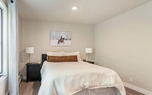 A bedroom with a bed, two bedside tables, a nightstand, a window with curtains, and a painting on the wall.