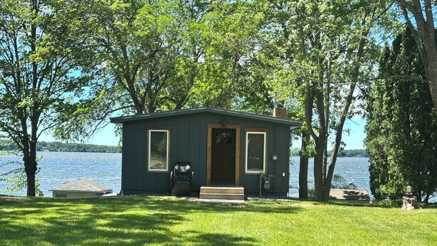 A small cabin with a door and windows, surrounded by trees and a lake.