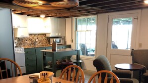 A dining area with a wooden table and chairs, a kitchen in the background, and a view through a window.