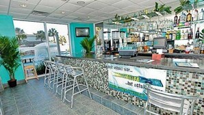 Zona de bar con salpicadero de azulejos, taburetes y mostrador con una computadora y varias botellas.