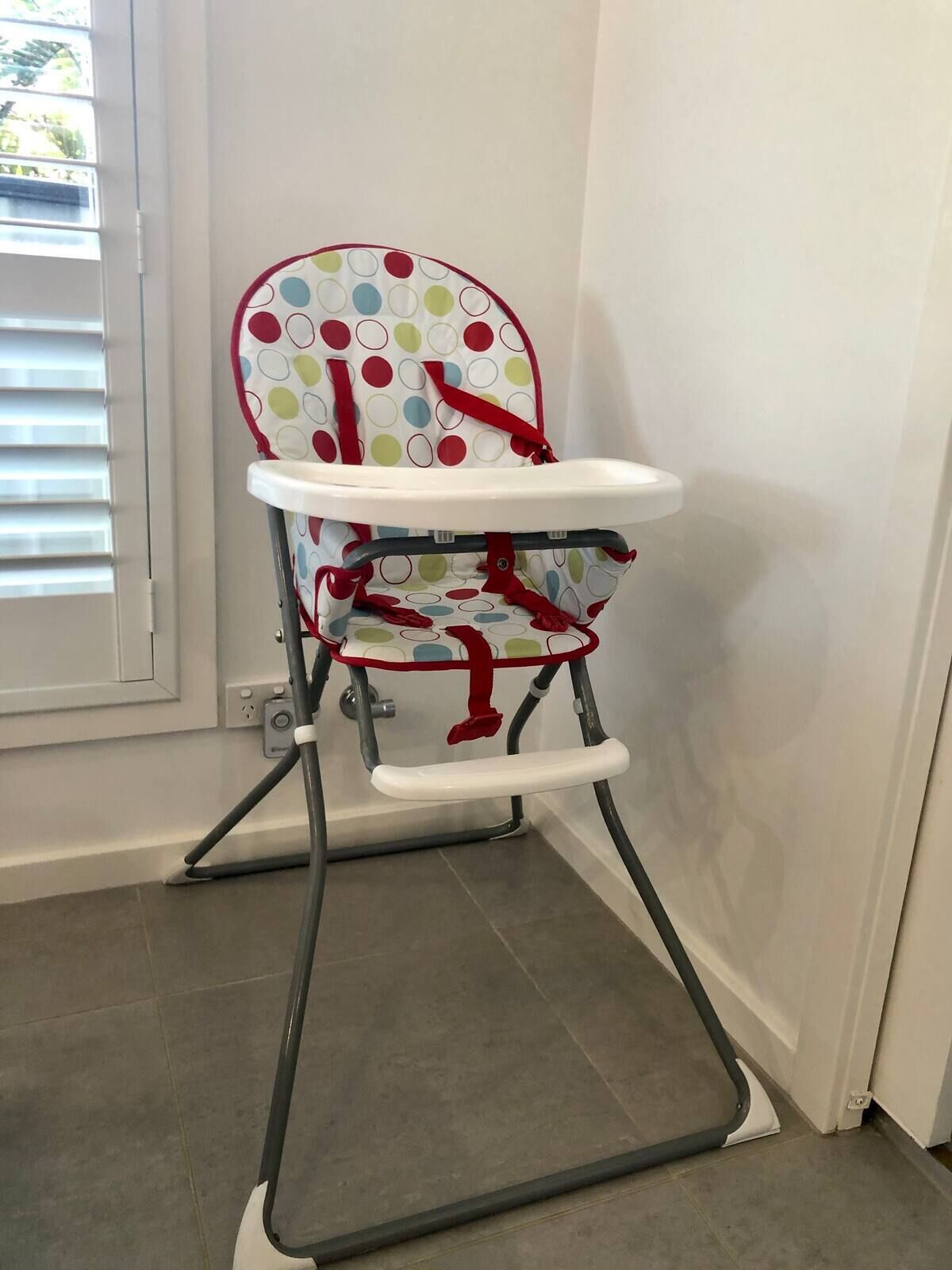 A high chair with a colorful polka dot pattern and red accents, positioned in a room with a white wall and a window with blinds.