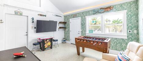 A room with a foosball table, a ping pong table, a TV, and a window with a view of greenery.