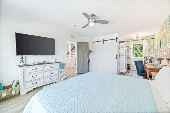 A bedroom with a large bed, a ceiling fan, a flat-screen TV, a dresser, and a door leading to another room.