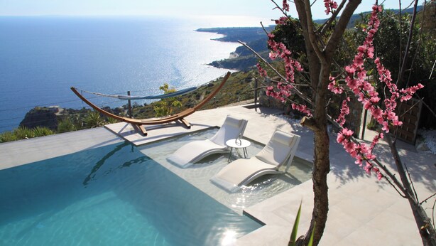 Una piscina con vista al océano, una hamaca y sillones de jardín blancos.