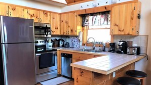 A modern kitchen with wooden cabinets, stainless steel appliances, a central island with stools, and a window with patterned curtains. - Welcome to LAZY DEER CABIN! (Piney Creek)