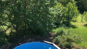 A circular swimming pool with a wooden deck surrounding it, set in a lush garden with trees and shrubs. - Gîte 12 per (Vers)