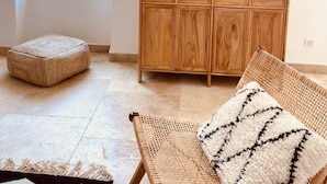 Una habitación con un ventanal grande, un aparador de madera con puertas de cristal, un otoman tejido, una silla tejida con un cojín estampado y un macramé colgado en la pared.