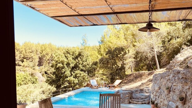 Zona de piscina al aire libre con una mesa y sillas de madera, rodeada de muros de piedra y plantas en macetas.