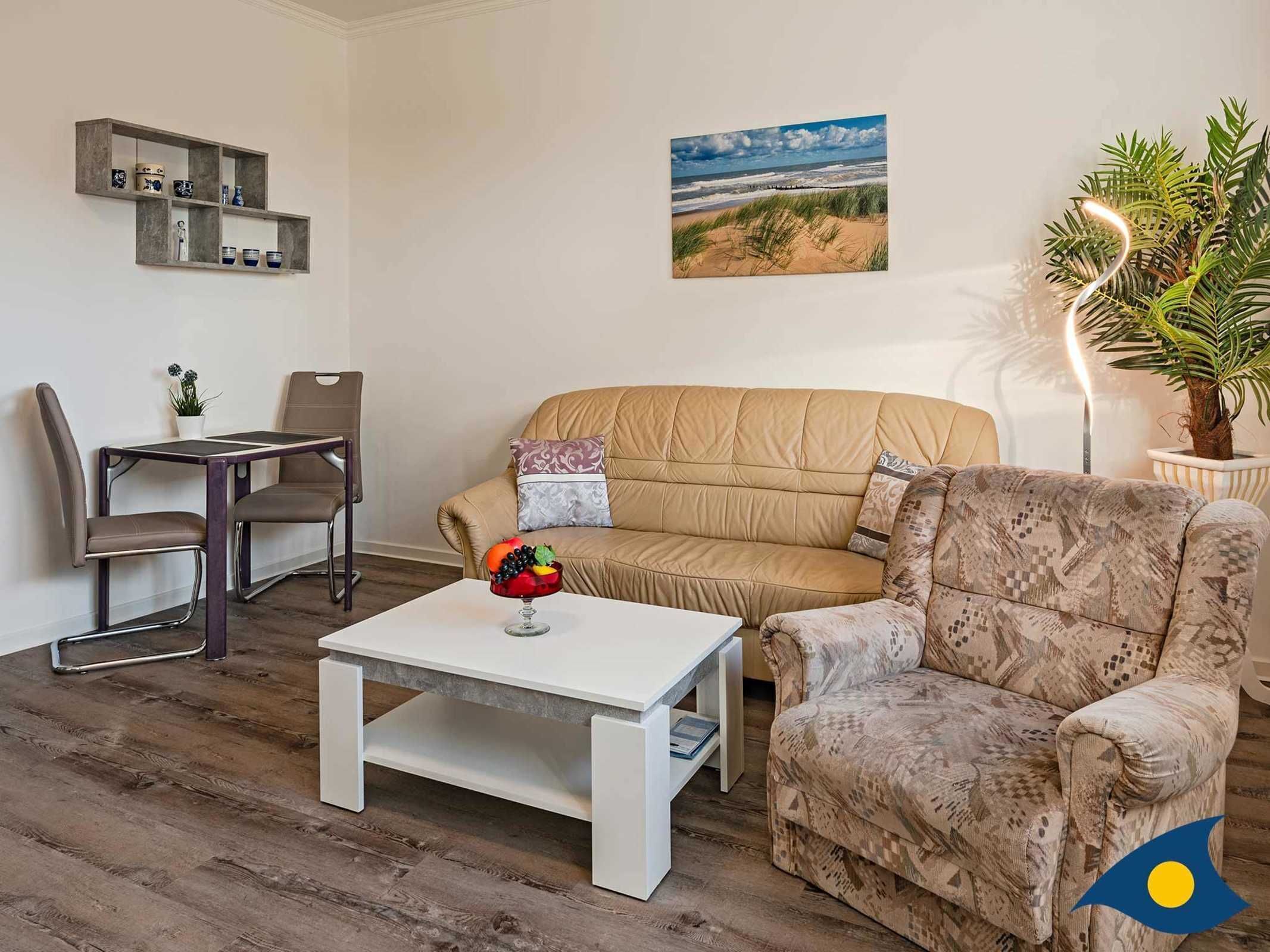 A living room with a beige sofa, patterned armchair, white coffee table, and a dining area with a table and chairs.