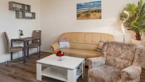 Un salon avec un canapé beige, un fauteuil à motifs, une table basse blanche et un coin repas comprenant une table et des chaises.