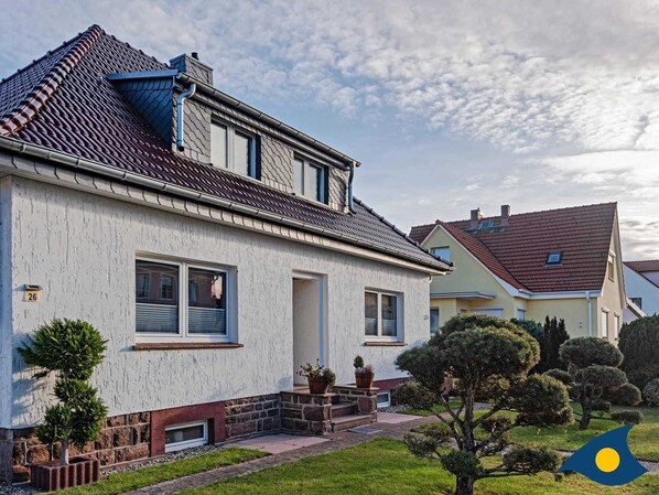 Une maison à deux étages, à l’extérieur blanc, avec un toit recouvert de tuiles rouges et un petit porche à l’avant, agrémenté de plantes en pot.