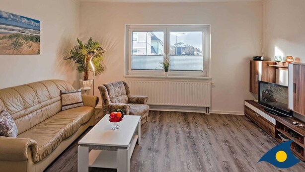 Un salon avec un canapé beige, un fauteuil plus petit, une table basse avec des fruits, un téléviseur posé sur un meuble en bois et une fenêtre avec des stores.