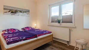 Une chambre avec un lit en bois, une fenêtre avec des stores, un radiateur et un petit tabouret en osier.