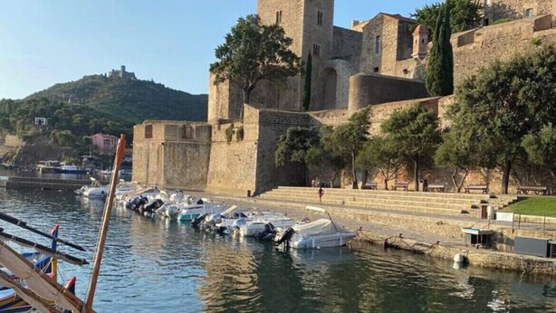A historic stone building with a tower, a dock with colorful boats, and a hilly landscape in the background. - 40m2 T2 apartment Small medieval village in the heart of the Roussillon vineyards (Baixas)